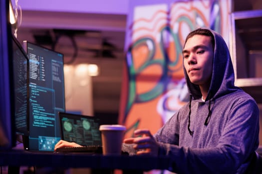 Asian hacker drinking coffee to go and hacking computer network system. Internet criminal in hood taking paper mug while coding illegal malicious software and breaking law