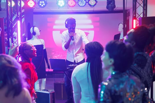 Young man dj mixing music using controller and singing in microphone on club stage with spotlights. African american musician entertaining clubbers crowd at party in nightclub