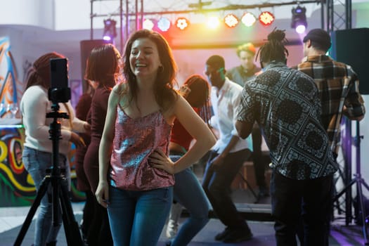 Cheerful smiling woman taking photo on smartphone while having fun in nightclub. Carefree laughing girl recording video using mobile phone on tripod while partying in club