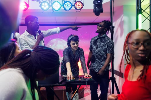 Musicians blending tracks with music mixer console and singing on stage in nightclub. African american men and caucasian woman band playing at disco party in crowded club