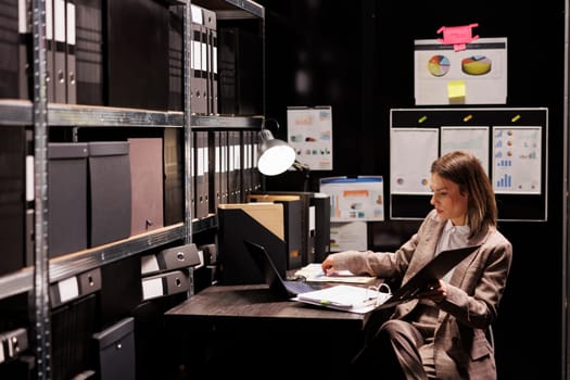 Manager analyzing administrative report, working late at night at bureaucracy record in arhive room. Corporate businesswoman reading accountancy documentation, trying to organizing files