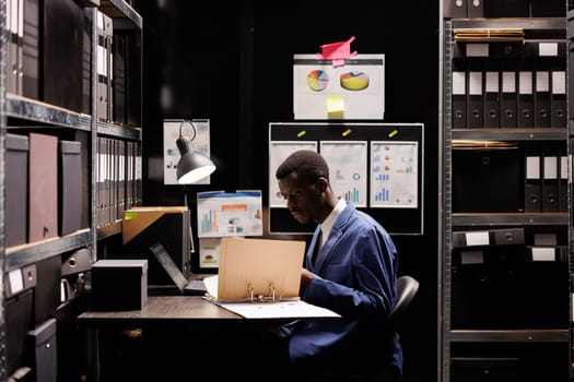 African american manager checking administrative files, analyzing bureaucracy record late at night in arhive room. Corporate worker in formal suit organizing accountancy documents. Depository concept