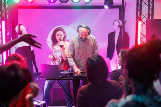 Woman singing and man dj mixing tracks while people partying on dancefloor in nightclub. People dancing and enjoying electronic music band show on stage with spotlights in club