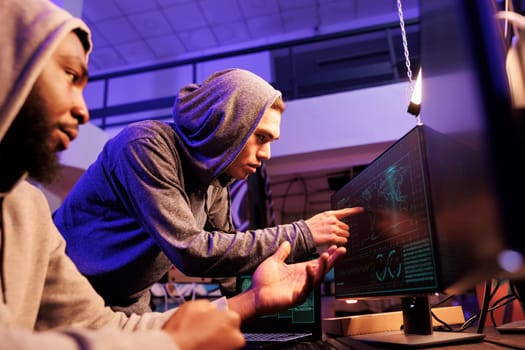 Hackers pointing at map location at computer screen while spying online. Two diverse young men in hoods using illegal spyware while doing criminal activity together at night