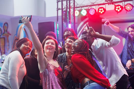 Happy diverse friends posing for smartphone selfie together while having fun at nightclub party. Cheerful young people taking photo on mobile phone while relaxing on club dancefloor