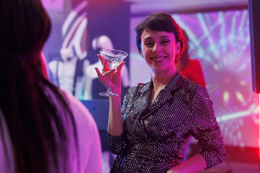 Smiling young woman drinking alcohol and partying on crowded nightclub dancefloor portrait. Cheerful happy clubber holding beverage glass and looking at camera at club discotheque