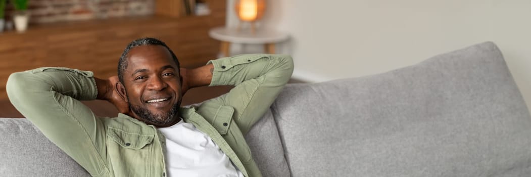 Cheerful middle aged african american man resting on sofa, enjoying free time alone, silence and comfort in cozy living room interior, close up, copy space. Relax, weekend break at home and lifestyle