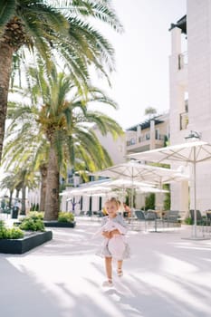 Little girl with a pink toy rabbit runs down the street past green palm trees. High quality photo