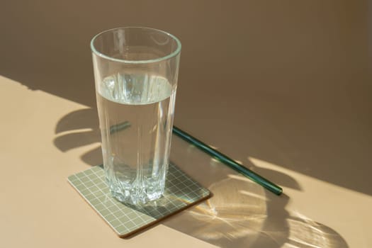 Glass of water in neutral beige background with shadows of leaves. Natural glass recyclable drinking straw. Zero waste sustainable lifestyle concept. Copy space