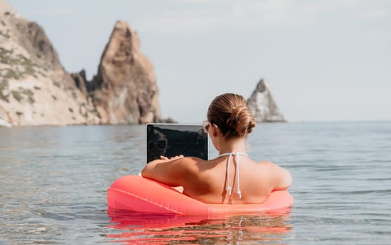 Woman freelancer works on laptop swimming in sea on pink inflatable ring. Pretty lady typing on computer while floating in the sea on inflatable donut at sunset. Freelance, remote work on vacation