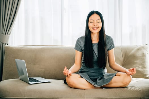 Peaceful Asian woman uses laptop on sofa meditating in lotus pose. Businesswoman seeks relaxation and balance embracing mindfulness while studying online. Smiling student in a serene office.