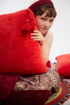 Portrait of Little girl in a stylized Tatar national costume having rest with a red pillow on a white background in the studio. Photo shoot of funny young teenager who is not a professional model