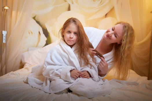 Mother and daughter happily relax and fun together on bed in bedroom. The concept of tenderness between mom and girl. Mom braids the girl's hair