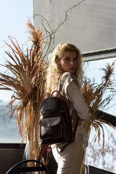 A woman holds brown leather backpack. Model wearing stylish knitted vest, white shirt and classic trousers.