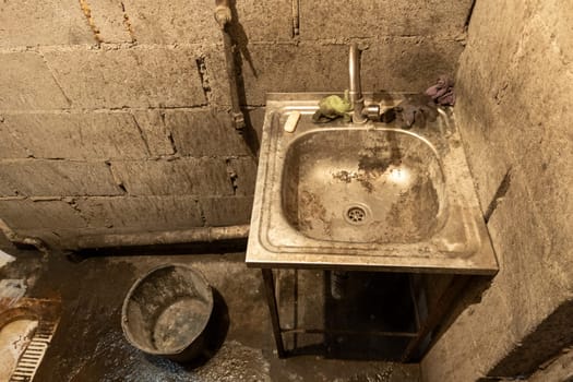 Dirty toilet room stainless steel sink with a bucket on the floor, uncoated concrete flooring and cinder brick walls. Developing country poorness.