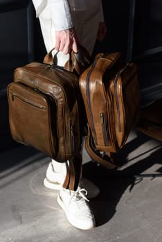 A woman holds two brown leather backpacks. Model wearing stylish knitted vest, white shirt and classic trousers.