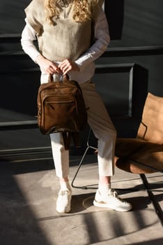 A woman holds brown leather backpack. Model wearing stylish knitted vest, white shirt and classic trousers.