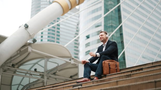 Smart business man wear headphone while sitting at stair in urban city and listen to relaxing music. Happy male leader using headset and moving to song in lively mood surrounded by skyscraper. Urbane.