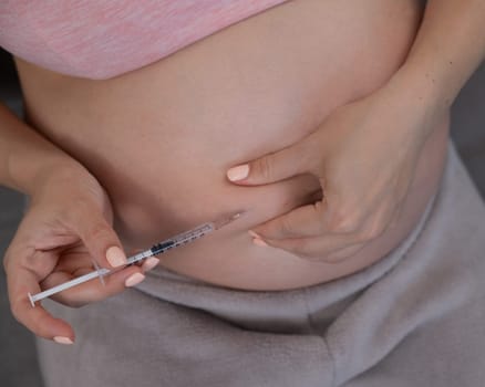 A pregnant woman puts an injection of insulin while sitting on the couch. Close up of the belly