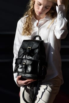 A woman holds black leather backpack. Model wearing stylish knitted vest, white shirt and classic trousers.