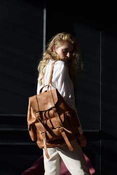 A woman holds brown leather backpack. Model wearing stylish knitted vest, white shirt and classic trousers.