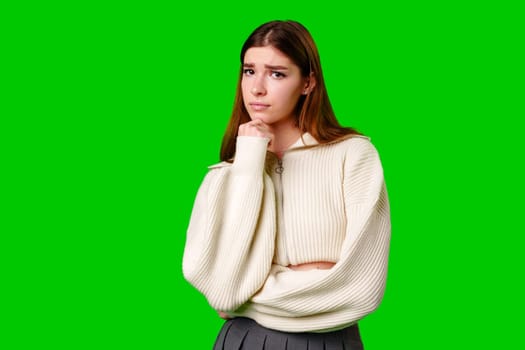 A young woman stands against a vivid green screen, her hand gently resting on her chin as she appears lost in thought, displaying a contemplative expression that suggests she is pondering a serious question or decision.