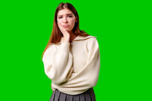 A young woman rests her cheek on her hand, conveying a look of boredom or disinterest. She is wearing a cozy white sweater and is positioned in front of a vivid green backdrop that contrasts with her neutral attire and indifferent expression.