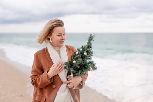 Blond woman Christmas sea. Christmas portrait of a happy woman walking along the beach and holding a Christmas tree in her hands. She is wearing a brown coat and a white suit