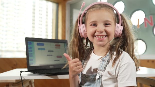 Closeup of caucasian girl smiling to camera while showing a thumb up gesture. Young child wearing headphone and casual dress standing while looking at camera with satisfy, happy, joyful. Erudition.