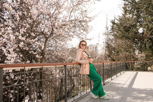 A woman is standing on a ledge with a cup of coffee in her hand. She is wearing sunglasses and a tan coat. The scene is set in a park with cherry blossoms in bloom. The woman is enjoying the view