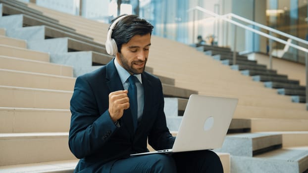 Confident smart business man celebrate successful business idea while listen music. Happy manager project working by using laptop while wearing headphone and suit. Investor sitting at stair. Exultant.