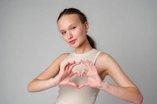 Young woman gesturing heart shape with hands on gray background in studio