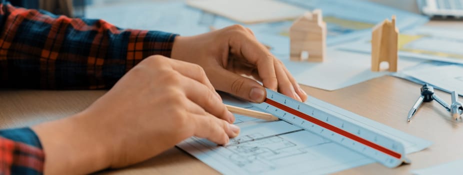 Closeup of architect engineer hand using ruler to mature and draw a blueprint on meeting table with wooden block, pencil and blueprint scatter around at architectural modern office. Delineation.