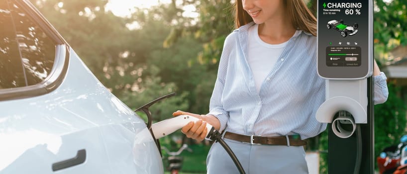 Young woman recharging battery for electric car during road trip travel EV car in natural forest or national park with sunrise lighting. Eco friendly travel during vacation and holiday. Panorama Exalt