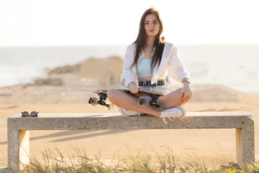 A woman is sitting on a bench with a skateboard in her hand. She is wearing a white shirt and blue shorts. The scene is set on a beach, with the ocean in the background