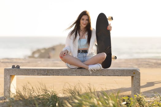 A woman is sitting on a bench with a skateboard in her hand. She is wearing a white shirt and blue shorts. The scene is set on a beach, with the ocean in the background