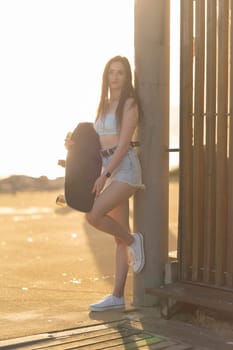 A woman is holding a skateboard and standing next to a fence. The image has a casual and relaxed vibe, with the woman posing for the camera in a beach setting
