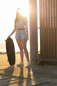 A woman is holding a skateboard and standing in front of a wooden fence. The image has a casual and relaxed vibe, with the woman posing for the camera