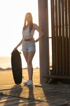 A woman is holding a skateboard and standing in front of a wooden fence. The image has a casual and relaxed vibe, with the woman posing for the camera in a beach setting