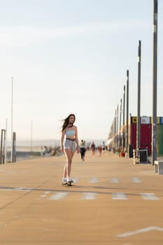 A woman is skateboarding down a sidewalk. She is wearing a blue tank top and shorts. There are other people walking around her, and a few benches are visible in the background