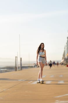 A woman is skateboarding on a sidewalk near the beach. She is wearing shorts and a tank top