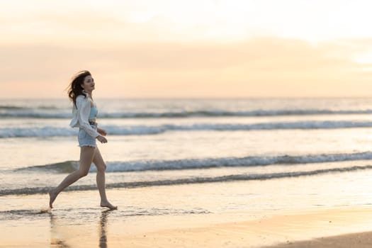 A woman is running on the beach at sunset. The sky is orange and pink, and the water is calm. The woman is wearing shorts and a white shirt