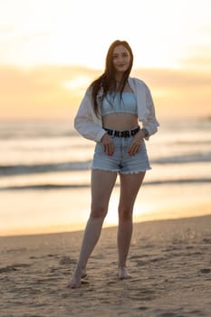 A woman is standing on the beach with her arms crossed and her hair wet. She is wearing a white shirt and blue shorts