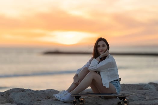 A woman is sitting on a skateboard on a beach. The sky is orange and the sun is setting