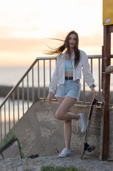 A woman is standing on a concrete ledge with a skateboard in her hand. She is wearing a white shirt and blue shorts. The image has a casual and relaxed vibe