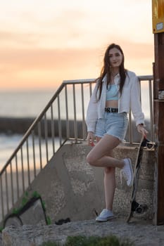 A woman is standing on a ledge with a skateboard in her hand. She is wearing a white jacket and blue shorts. The sky is orange and the water is in the background