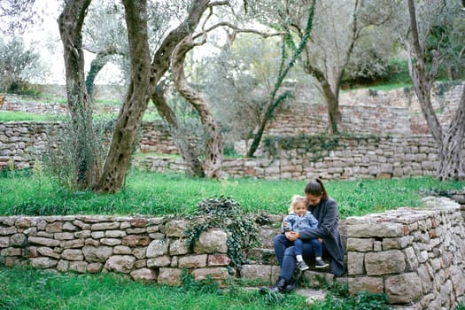Little girl sits on her mother lap on the steps of a stone fence in the park. High quality photo