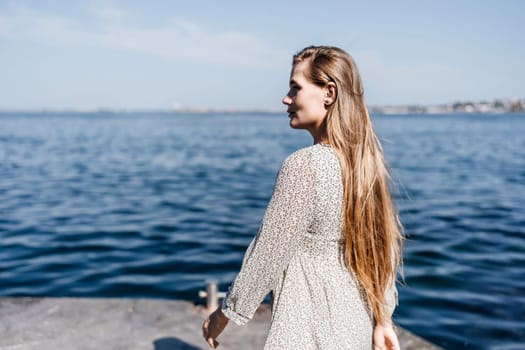 A woman is walking on the beach wearing a dress. The water is calm and the sky is clear