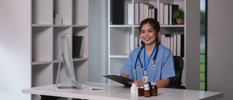 A woman in a blue scrubs is sitting at a desk with a computer and a clipboard. She is smiling and she is happy