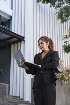 A woman in a business suit is holding a laptop and a folder. She is focused on her work
